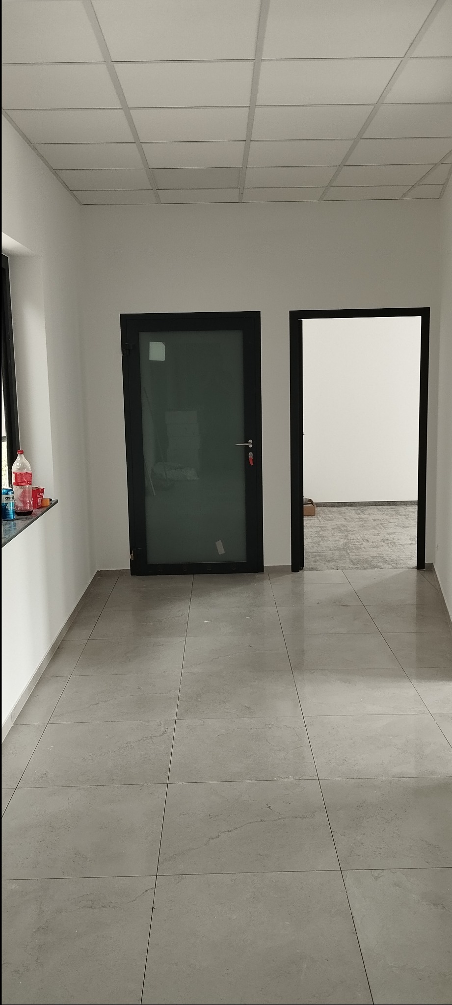Wnętrze z szarą, kwadratową podłogą z płytek, dwoje czarnych drzwi i białymi ścianami. Widoczne butelki na parapecie. Sufit kasetonowy.