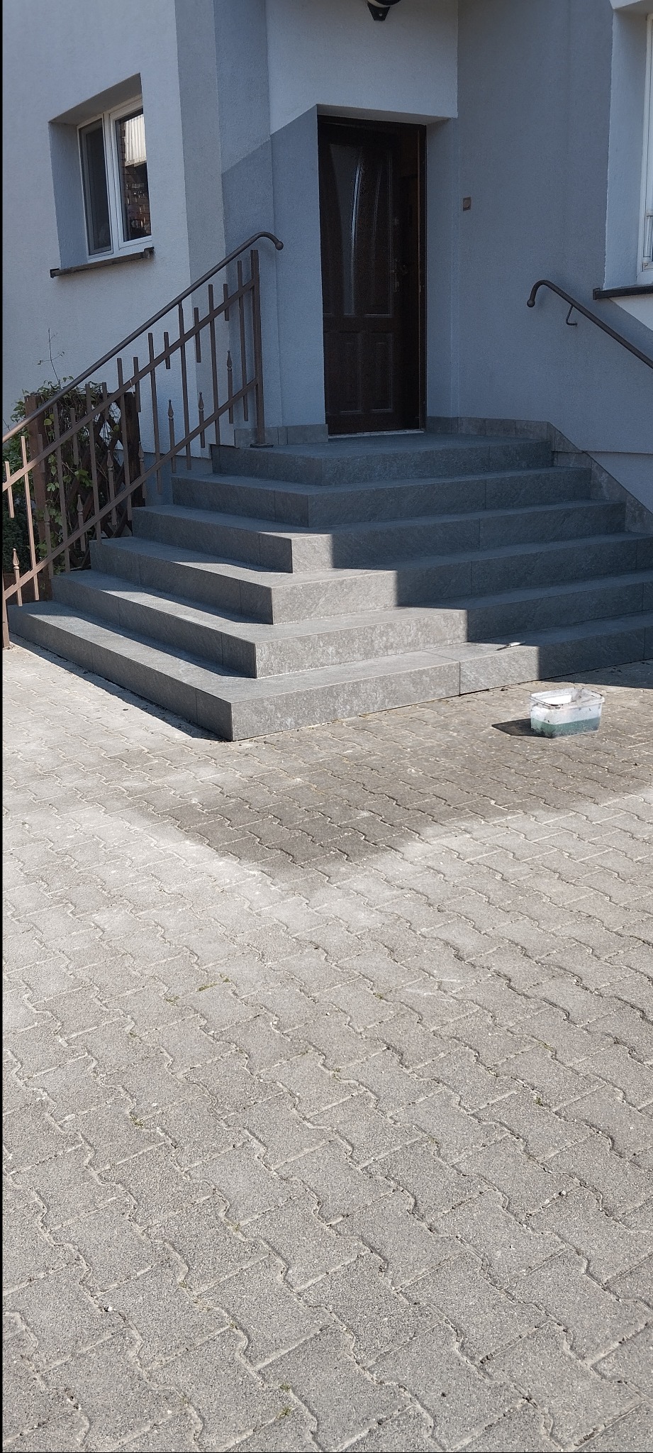 Schody wejściowe wyłożone szarą płytką, z metalową barierką. Widoczna kostka brukowa i wiadro z resztkami farby. Ujęcie z dołu, w dziennym świetle.