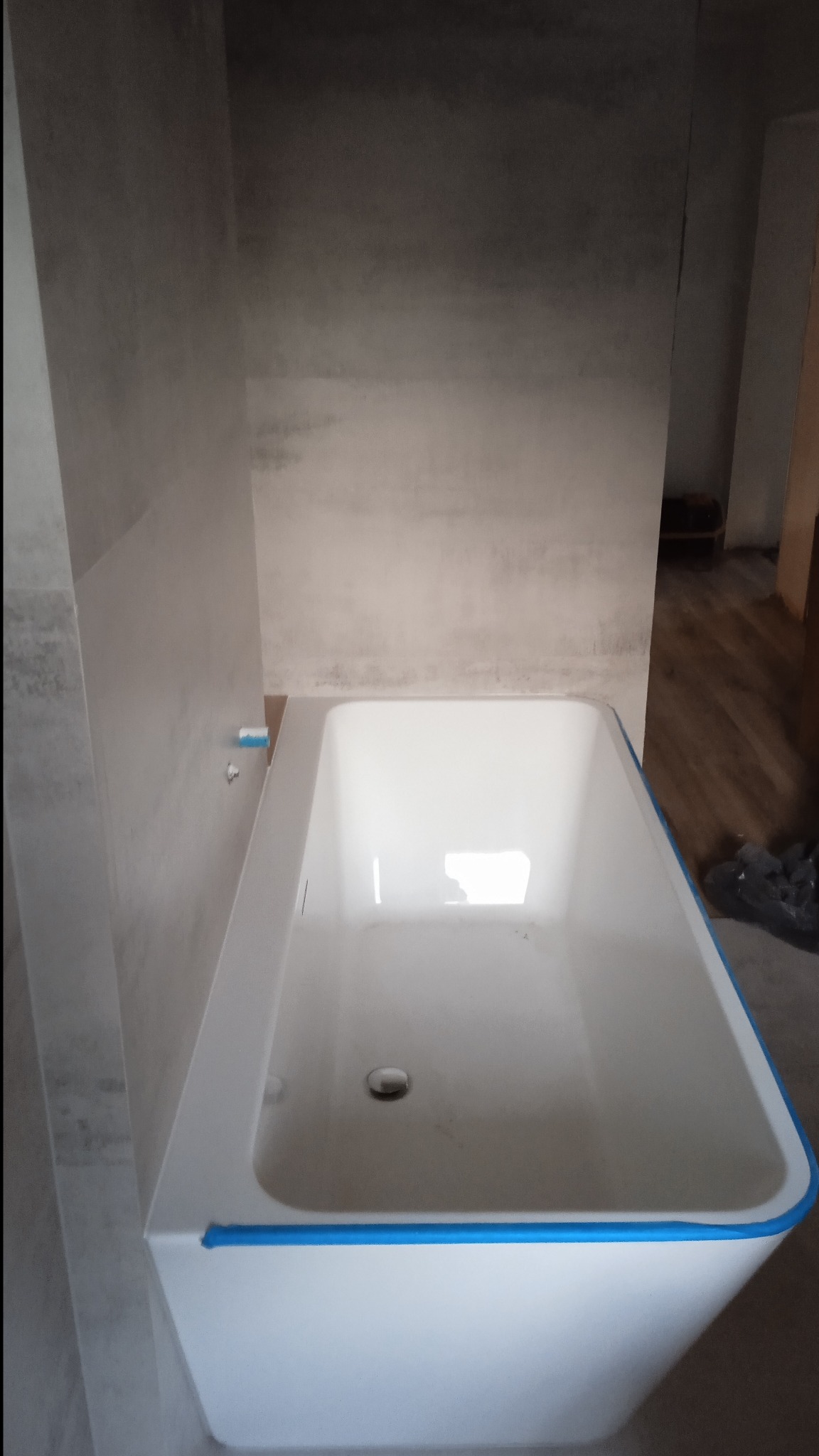 Biała wanna z niebieską taśmą zabezpieczającą krawędzie, zamontowana w surowej łazience z szarymi płytkami imitującymi beton. Widoczne elementy instalacji.