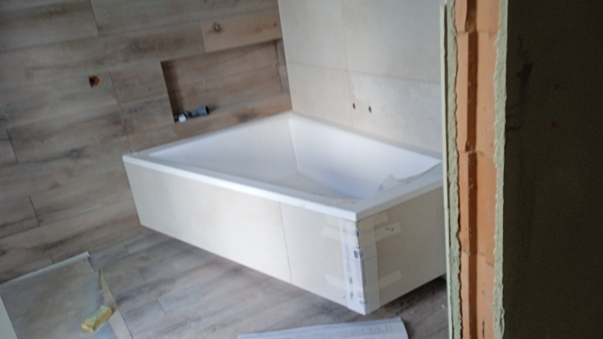 Wanna obudowana płytami w łazience z drewnopodobnymi płytkami na ścianach i podłodze. Widoczny fragment ościeżnicy.