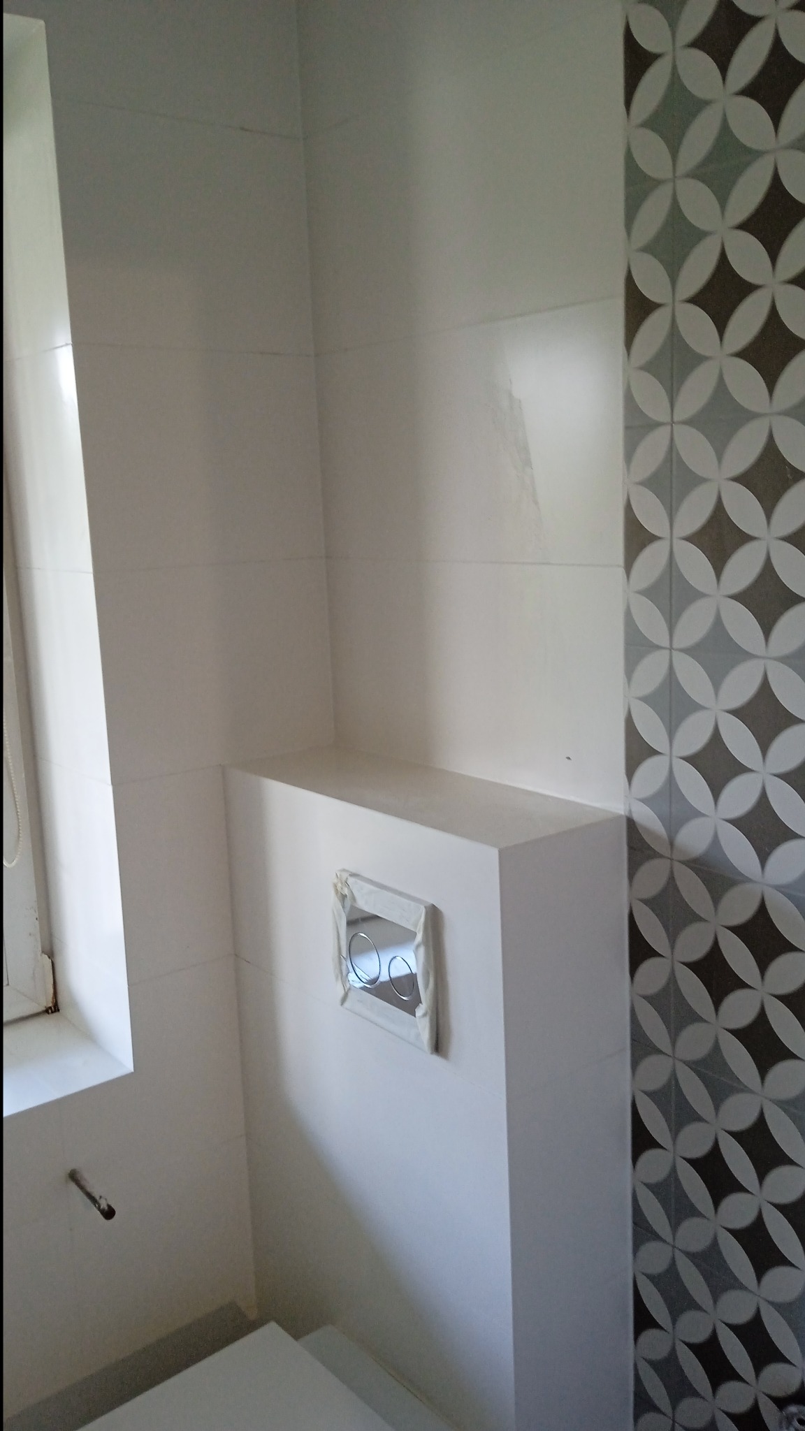 Biała łazienka po remoncie: płytki z geometrycznym wzorem, zabudowa geberitu z przyciskiem spłukującym, okno. Minimalistyczny design, czyste linie, jasne wnętrze.