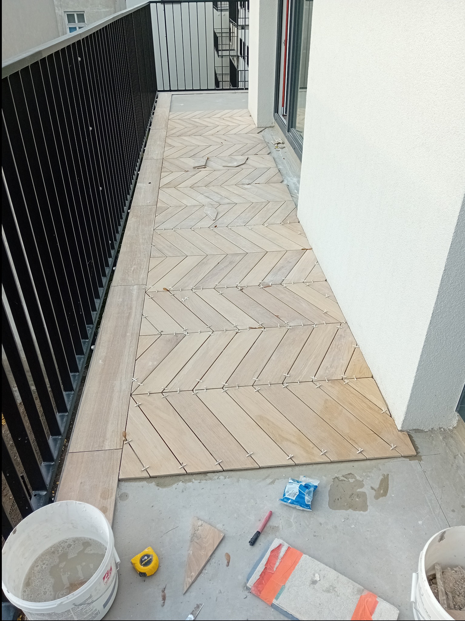 Układanie drewnopodobnych płytek w jodełkę na balkonie. Widoczne krzyżyki dystansowe, wiadro z zaprawą i narzędzia glazurnicze. Częściowo ukończona realizacja.