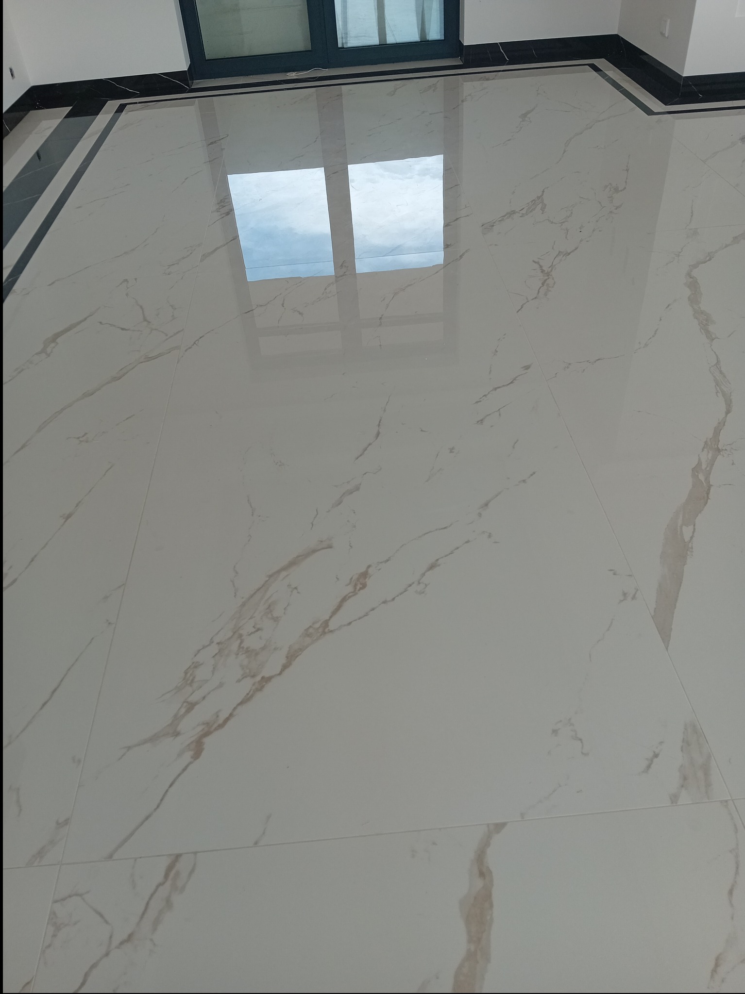Błyszcząca, biała podłoga z marmurowym wzorem i czarnymi listwami przypodłogowymi, odbijająca okno, świeżo po ułożeniu, z widocznymi fugami.