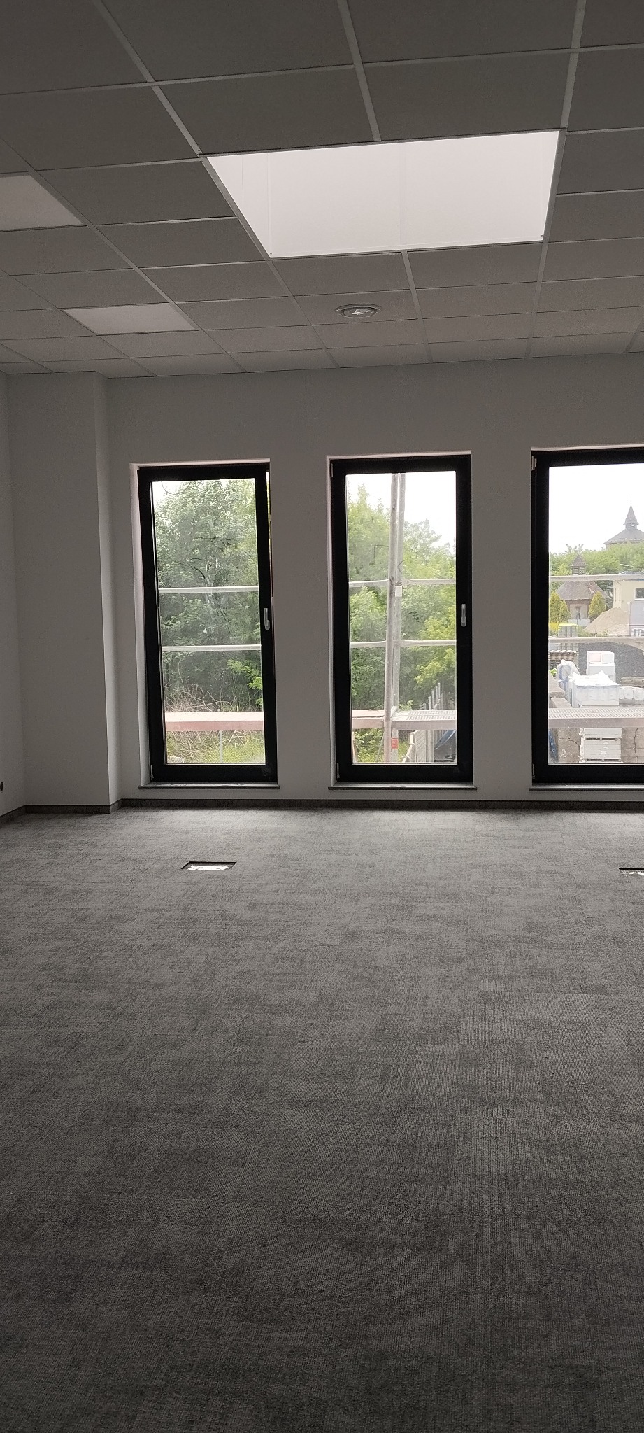 Wnętrze biura po remoncie: szara wykładzina dywanowa, trzy okna z czarnymi ramami, podwieszany sufit z oświetleniem. Widok na zieleń za oknami.