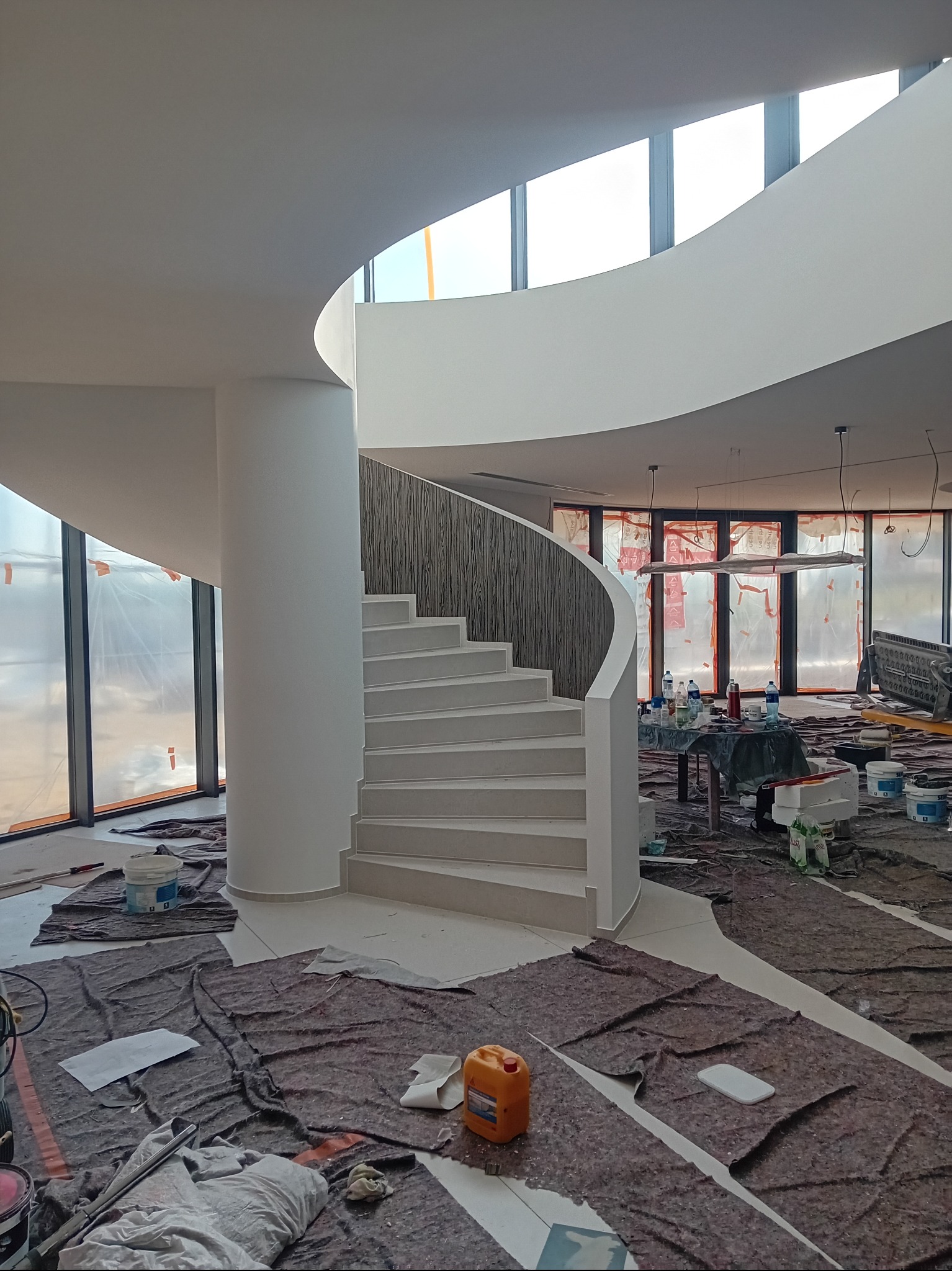 Wnętrze w trakcie malowania: spiralne schody z drewnianą balustradą, zabezpieczone podłogi, okna z folią ochronną. Widoczne puszki z farbą i narzędzia malarskie.