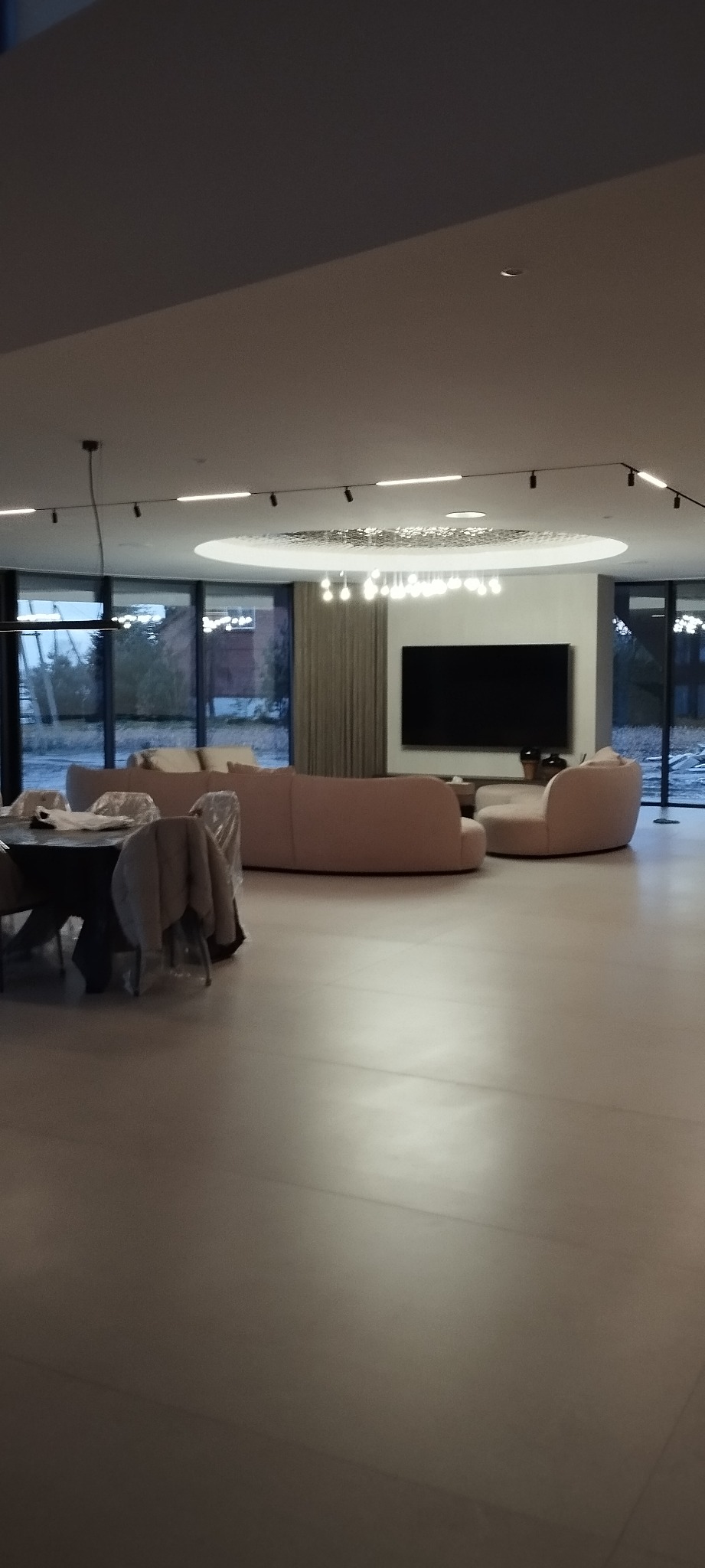 Nowoczesne wnętrze z dużym, jasnym salonem. Okrągły stół z krzesłami, za nim zaokrąglona sofa i telewizor na ścianie. Duże okna wpuszczają naturalne światło.