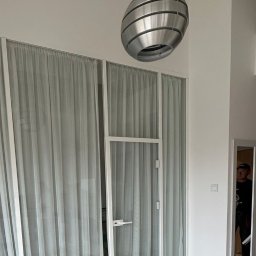 J&A MANAGER GROUP SPÓŁKA Z OGRANICZONĄ ODPOWIEDZIALNOŚCIĄ - Nowoczesne, białe drzwi szklane z zasłonami w minimalistycznym wnętrzu, oświetlone designerską lampą wiszącą. Widoczne detale wykończenia i odbicie osoby w lustrze.