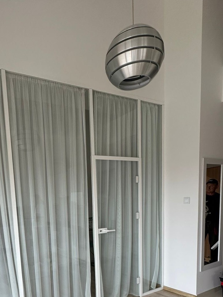 Nowoczesne, białe drzwi szklane z zasłonami w minimalistycznym wnętrzu, oświetlone designerską lampą wiszącą. Widoczne detale wykończenia i odbicie osoby w lustrze.