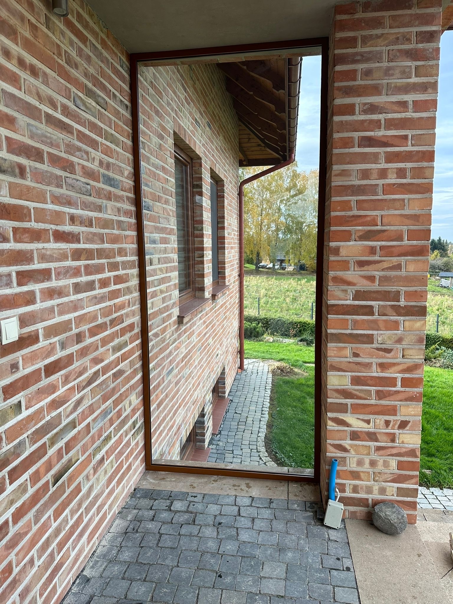 Szklana ściana na balkonie z cegły w Rzeszowie, widok na ogród. Ciemna rama okienna, szara kostka brukowa na podłodze balkonu, ceglana elewacja budynku.