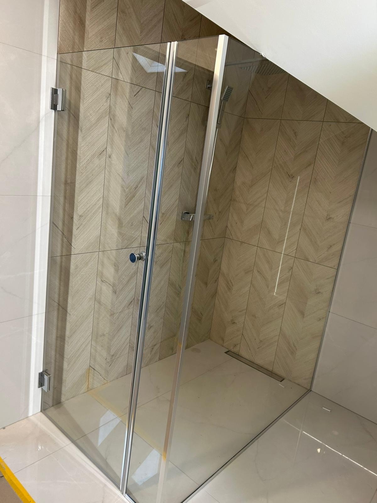 Kabina prysznicowa ze szklanymi drzwiami, wykończona płytkami w odcieniach beżu i bieli, z widocznym odpływem liniowym. Ujęcie z niskiego kąta.