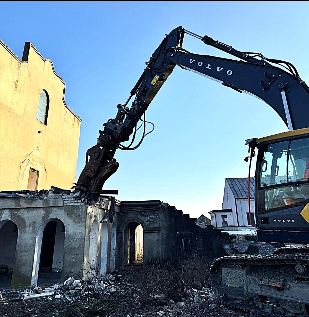 Częściowy demontaż budynku z łukowymi oknami przy użyciu koparki Volvo z hydraulicznymi nożycami. W tle żółta ściana budynku i błękitne niebo.