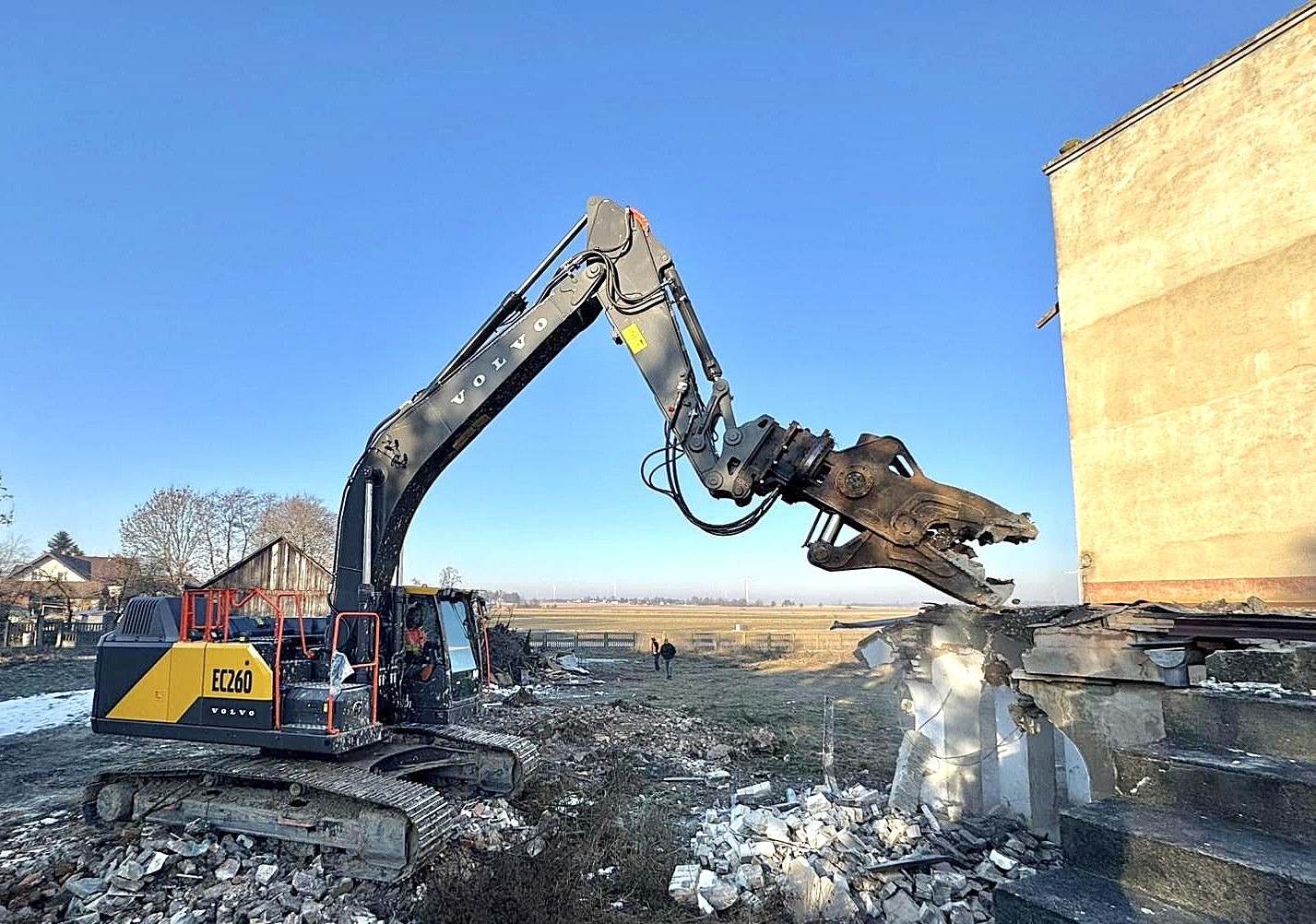 Żółto-czarna koparka Volvo EC260 z hydraulicznymi nożycami do betonu rozbiera fragment budynku na tle błękitnego nieba i krajobrazu z polami.