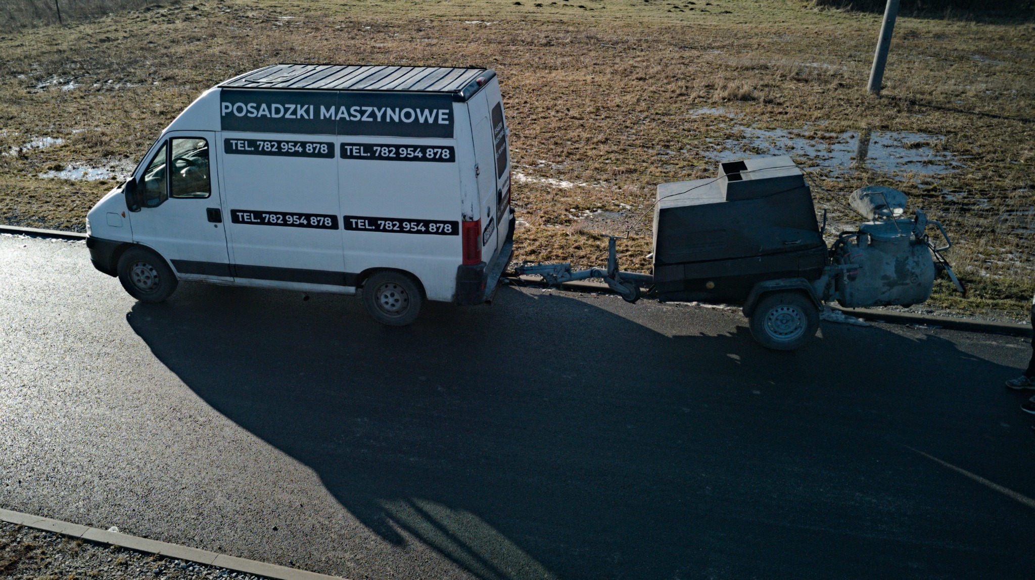 Biały samochód dostawczy z przyczepą i napisem 'Posadzki Maszynowe' na tle pola i asfaltowej drogi. Widok z góry, ostre światło słoneczne.