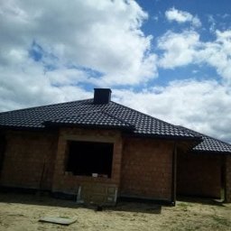 Usługi ciesielsko dekarskie - Dom w trakcie budowy z nowym, czarnym dachem z dachówki ceramicznej. Widoczne niezabudowane okna i komin. Błękitne niebo z chmurami w tle.