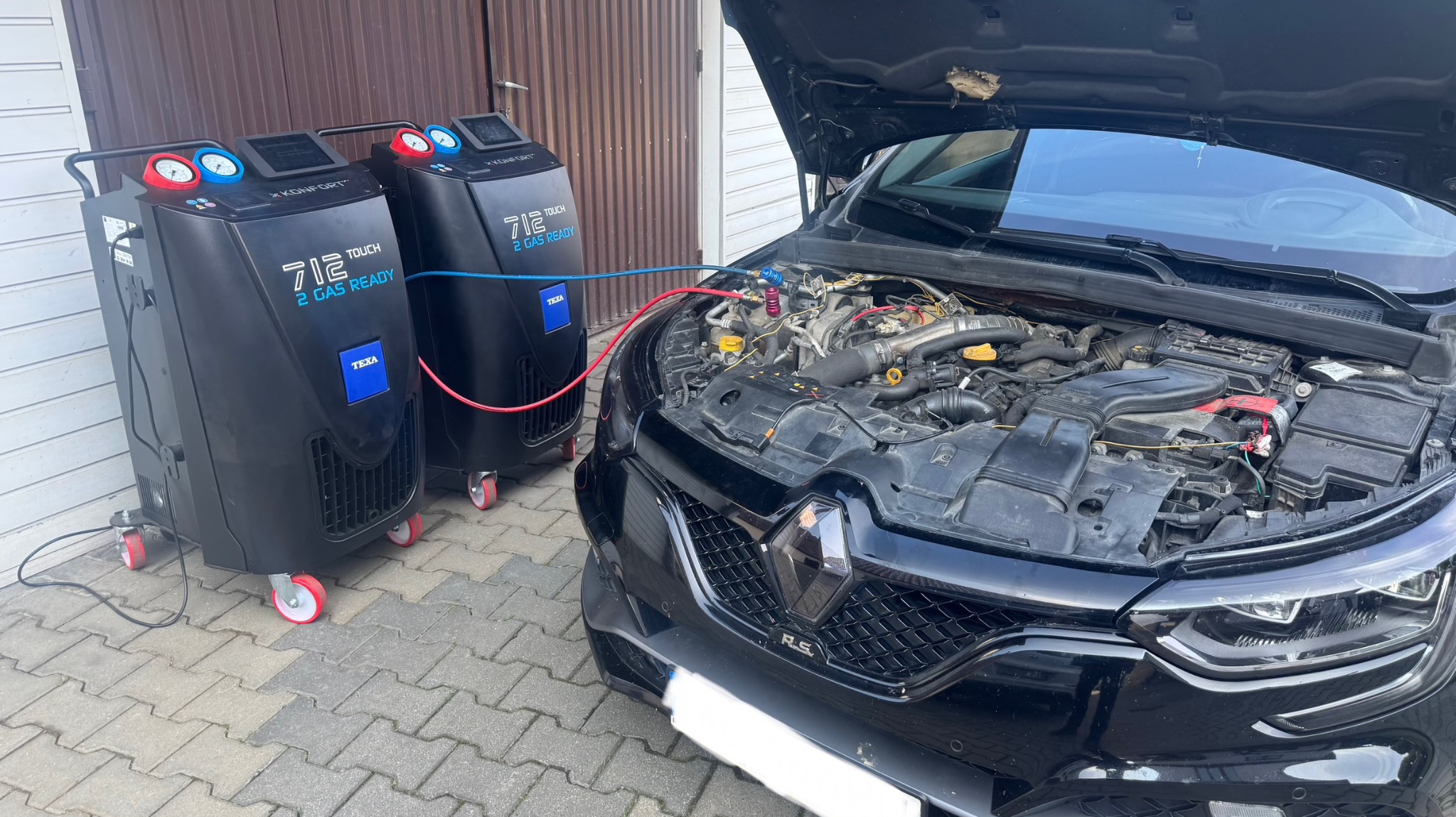 Czarny samochód z otwartą maską, podłączony do dwóch urządzeń TEXA 712 Touch 2 Gas Ready, prawdopodobnie podczas serwisu klimatyzacji. Widoczne przewody i silnik.
