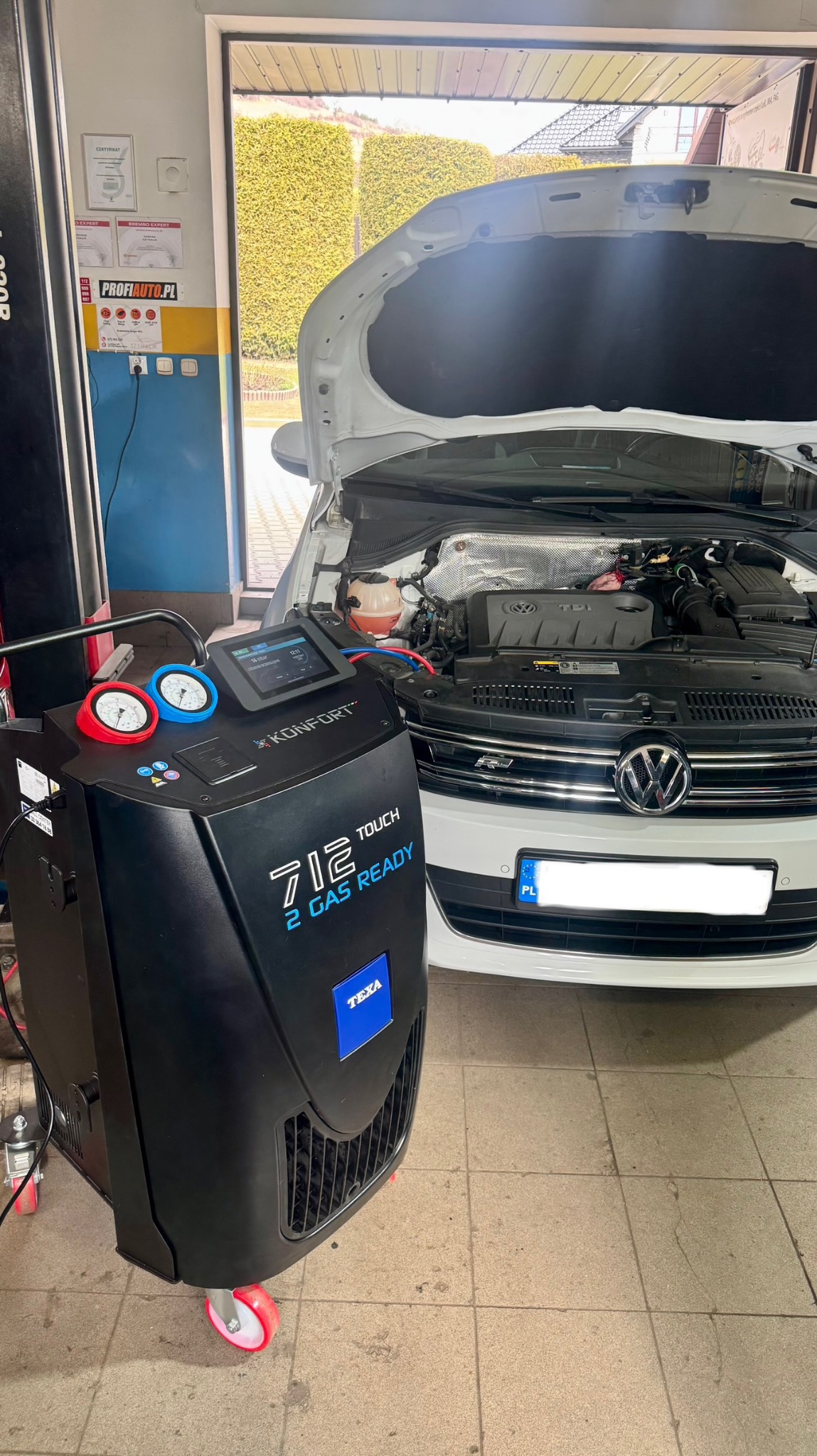 Biały samochód z otwartą maską w warsztacie, podłączony do czarnego urządzenia Texa do serwisowania klimatyzacji. Widoczne manometry i ekran dotykowy.