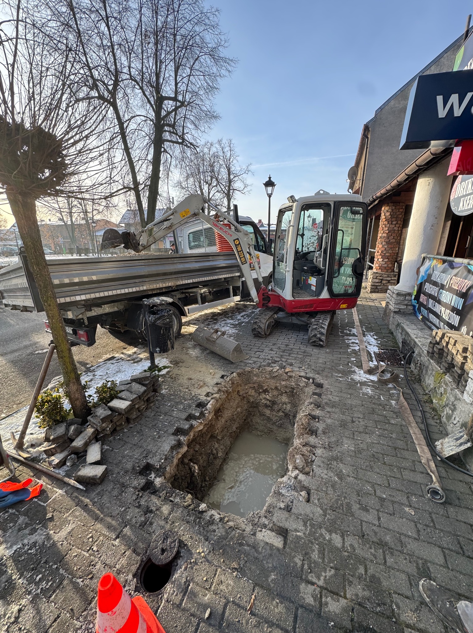 Mini koparka i wywrotka przy wykopie w kostce brukowej. Prace ziemne w otoczeniu miejskiej zabudowy, widoczny fragment chodnika i budynki w tle.