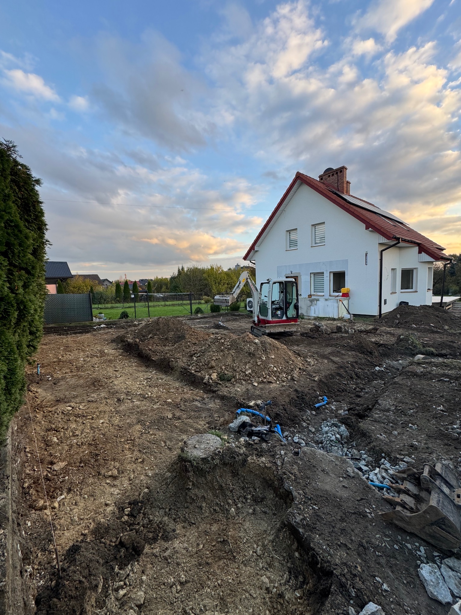 Mini koparka przy wykopie fundamentów domu jednorodzinnego. Widoczne wytyczone rowy, przygotowany teren pod budowę, dom w tle i pochmurne niebo.