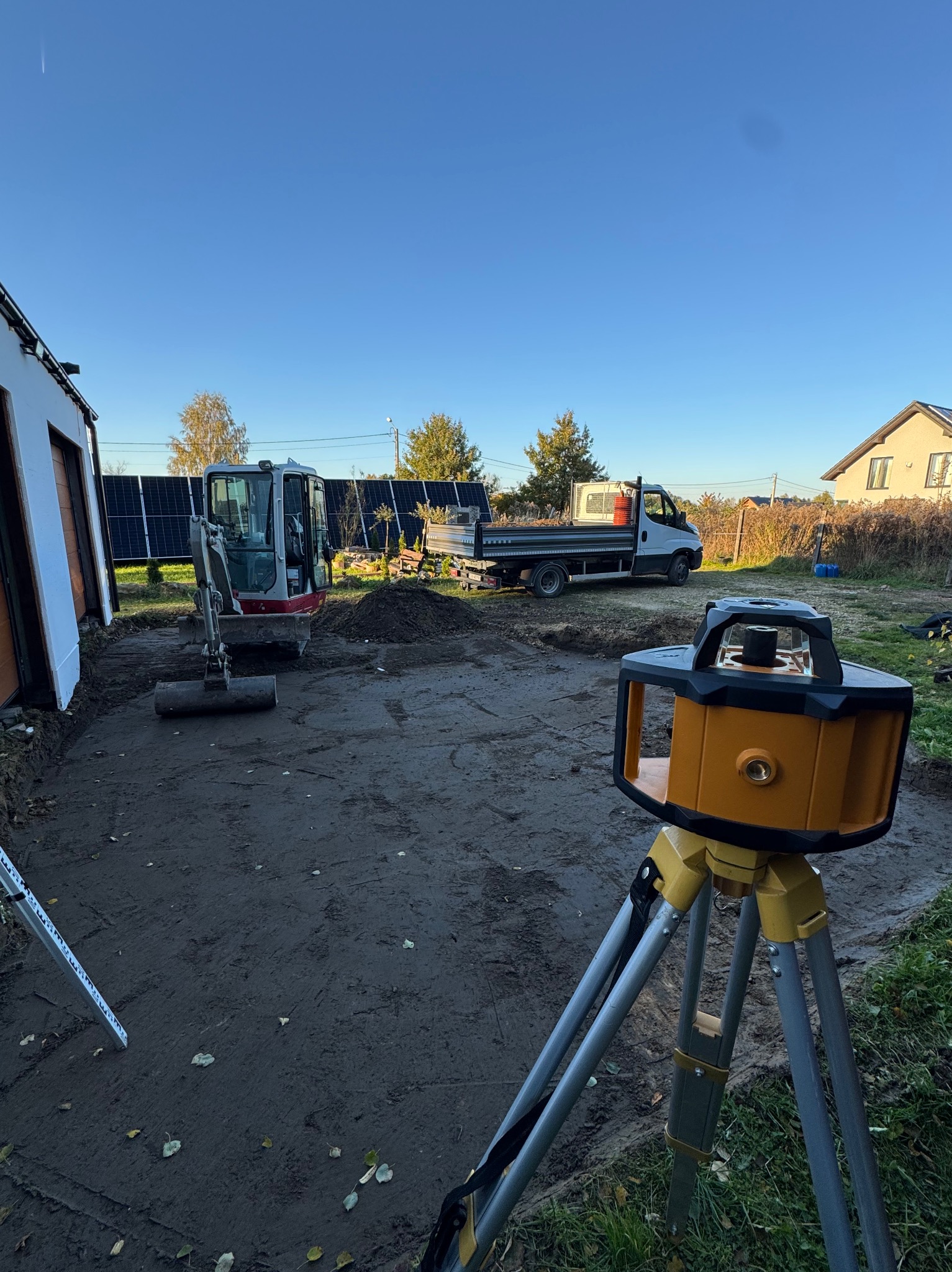 Mini koparka na placu budowy, obok ustawiony niwelator laserowy. W tle widoczny samochód dostawczy i budynek z panelami słonecznymi. Błękitne niebo.