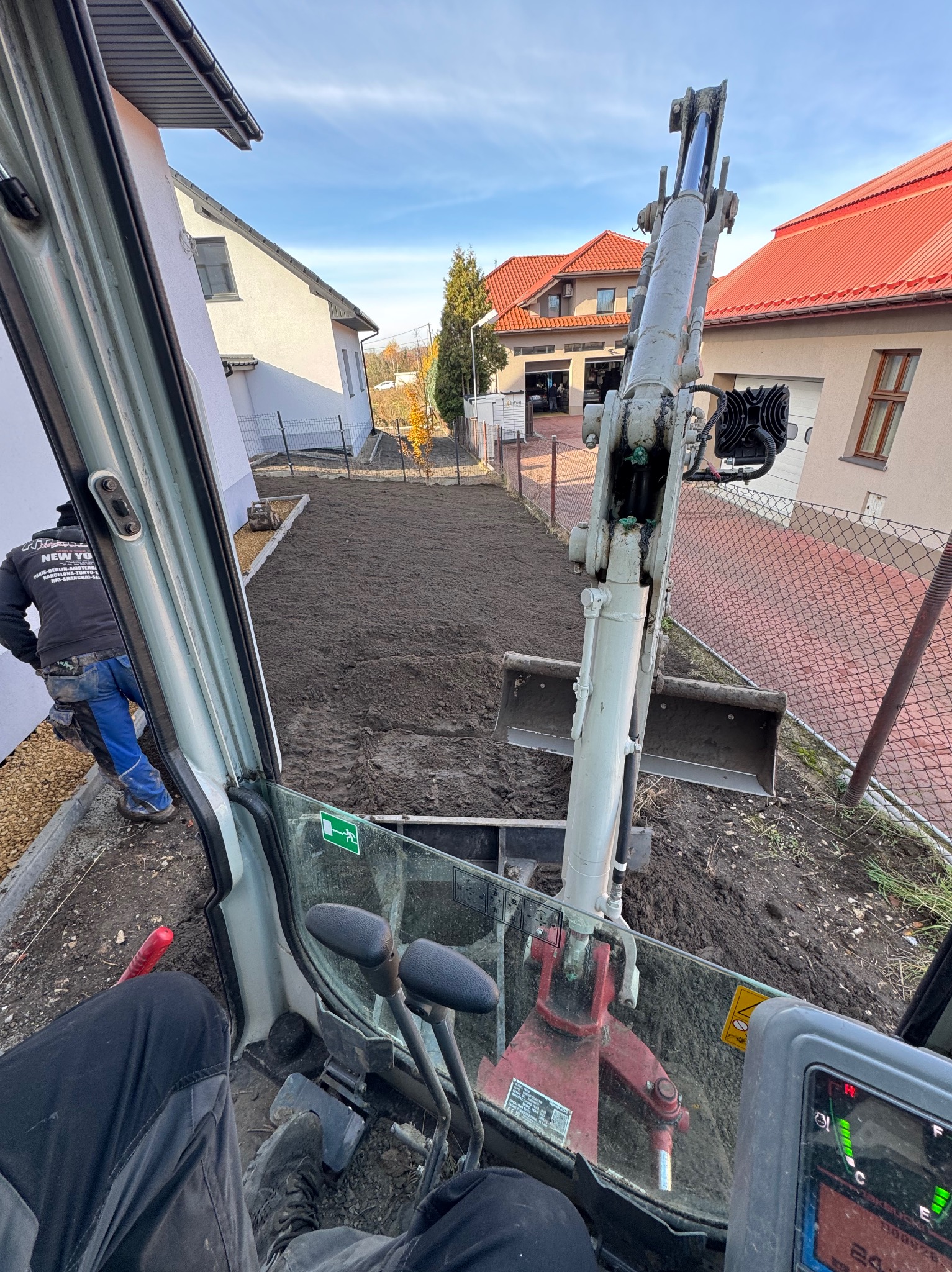 Widok z kabiny minikoparki na wyrównany teren działki z widocznymi budynkami w tle, częściowo ogrodzony siatką. Prace ziemne w toku.