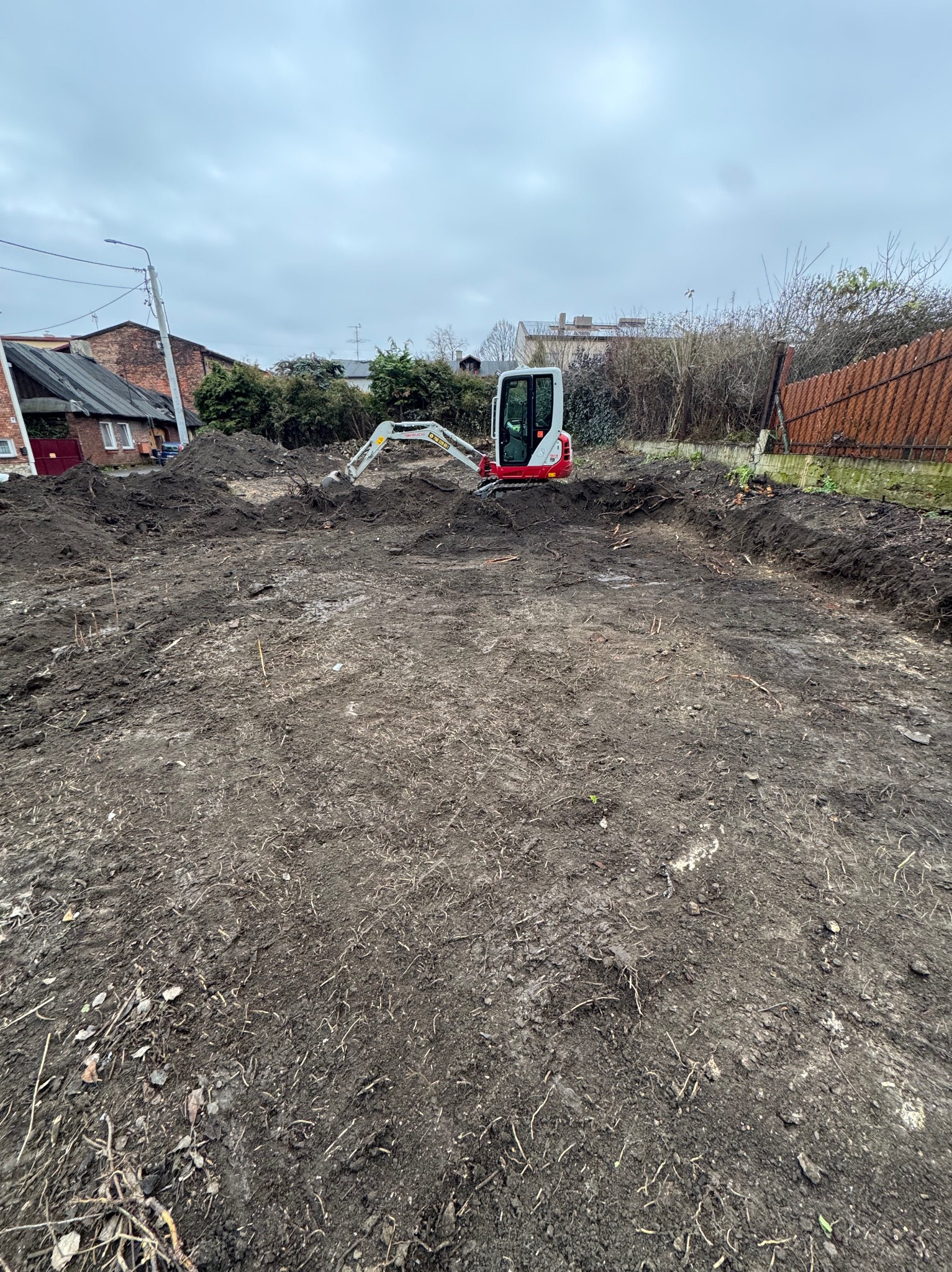 Mini koparka na placu budowy, świeżo po wykopie. Widoczne ślady opon na ziemi i przygotowany teren pod dalsze prace. W tle budynki mieszkalne i pochmurne niebo.