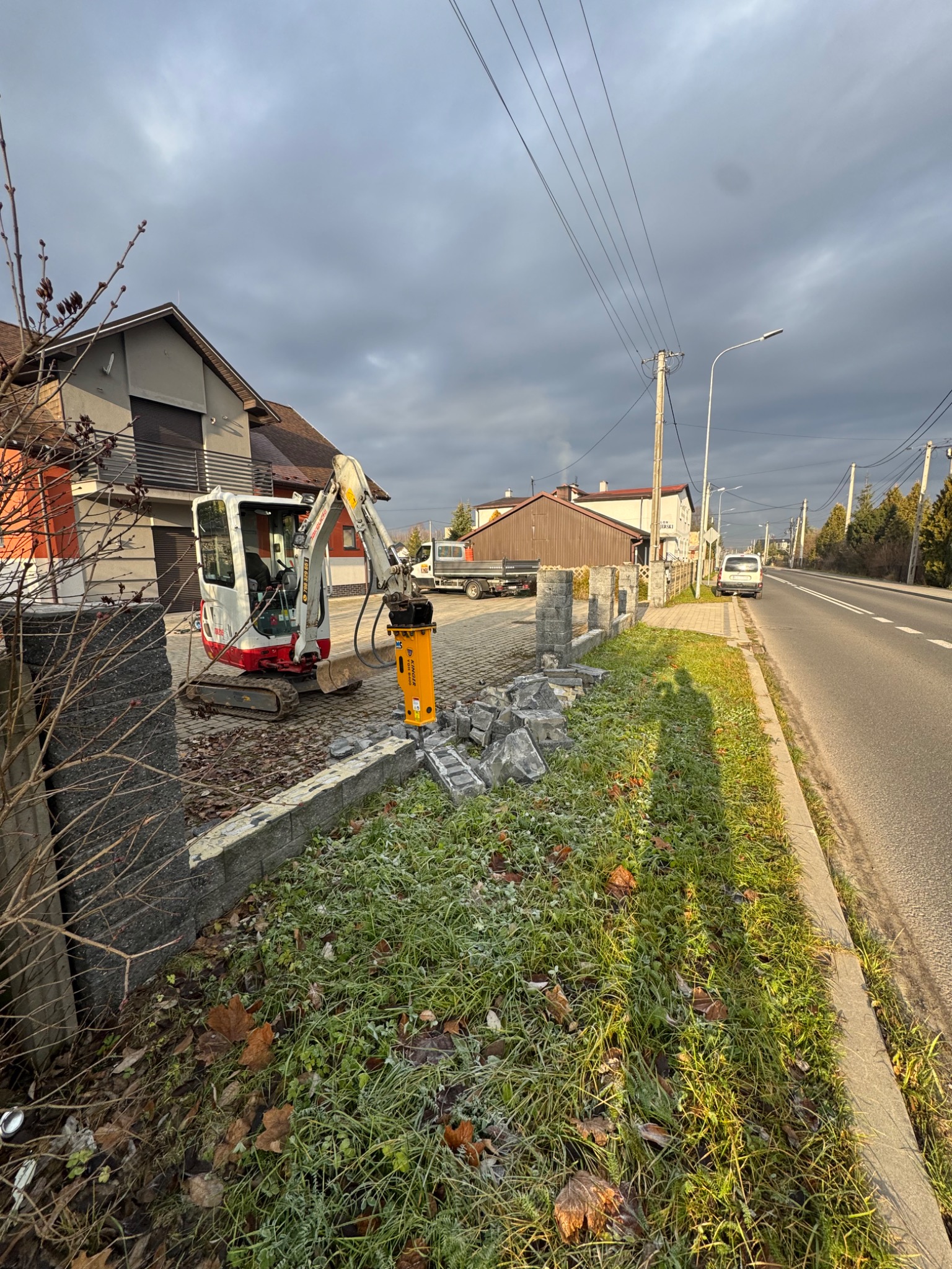 Mini koparka z młotem pneumatycznym kruszy betonowe bloczki przy drodze. Widoczne fragmenty zniszczonego murku i trawa z szronem. Pochmurne niebo.