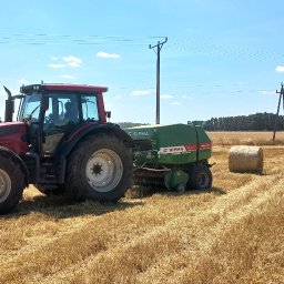 LasRol Jaworscy Mateusz Jaworski - Czerwony traktor z zieloną prasą zwijającą na polu po żniwach, z balotami słomy i słupami energetycznymi w tle, pod błękitnym niebem.
