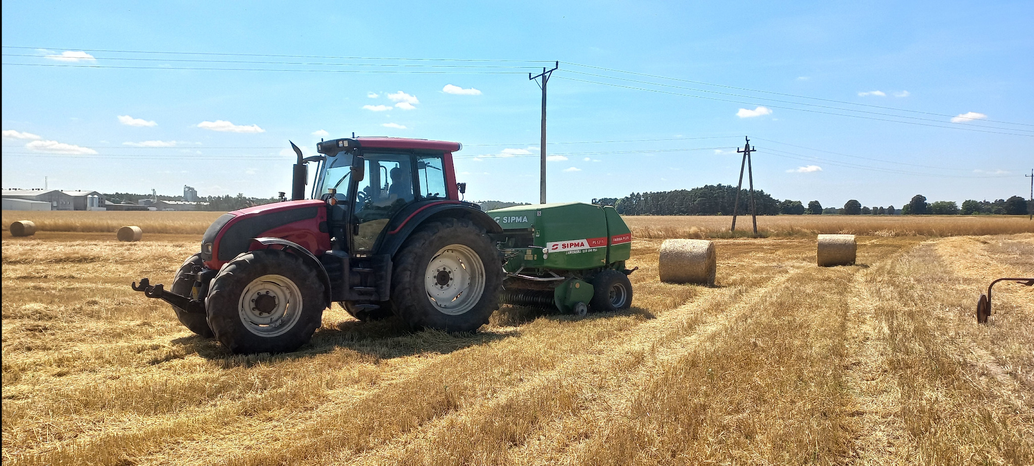 Czerwony traktor z zieloną prasą zwijającą na polu po żniwach, z balotami słomy i słupami energetycznymi w tle, pod błękitnym niebem.
