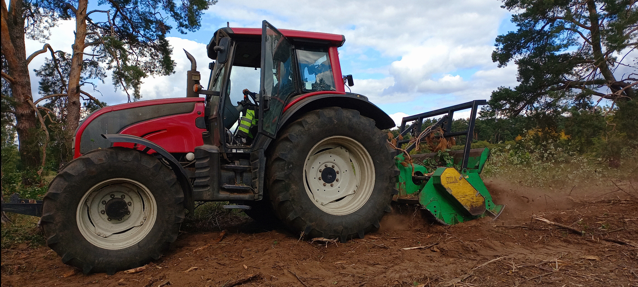Czerwony traktor z mulczerem leśnym w akcji, rozdrabniający gałęzie na zrębie. Widoczne drzewa w tle i pył unoszący się podczas pracy.