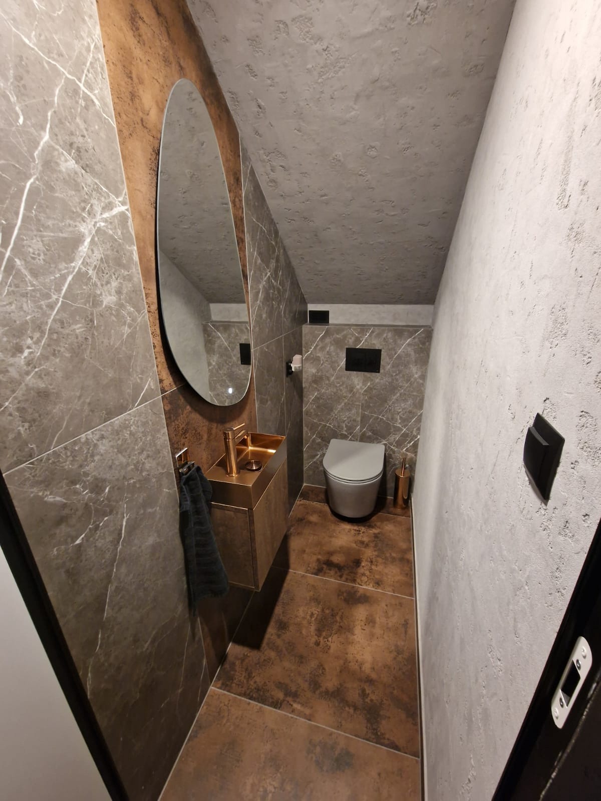 Nowoczesna łazienka z marmurowymi płytkami, złotą umywalką i owalnym lustrem. Widoczna toaleta i teksturowana ściana. Minimalistyczny design z akcentami luksusu.