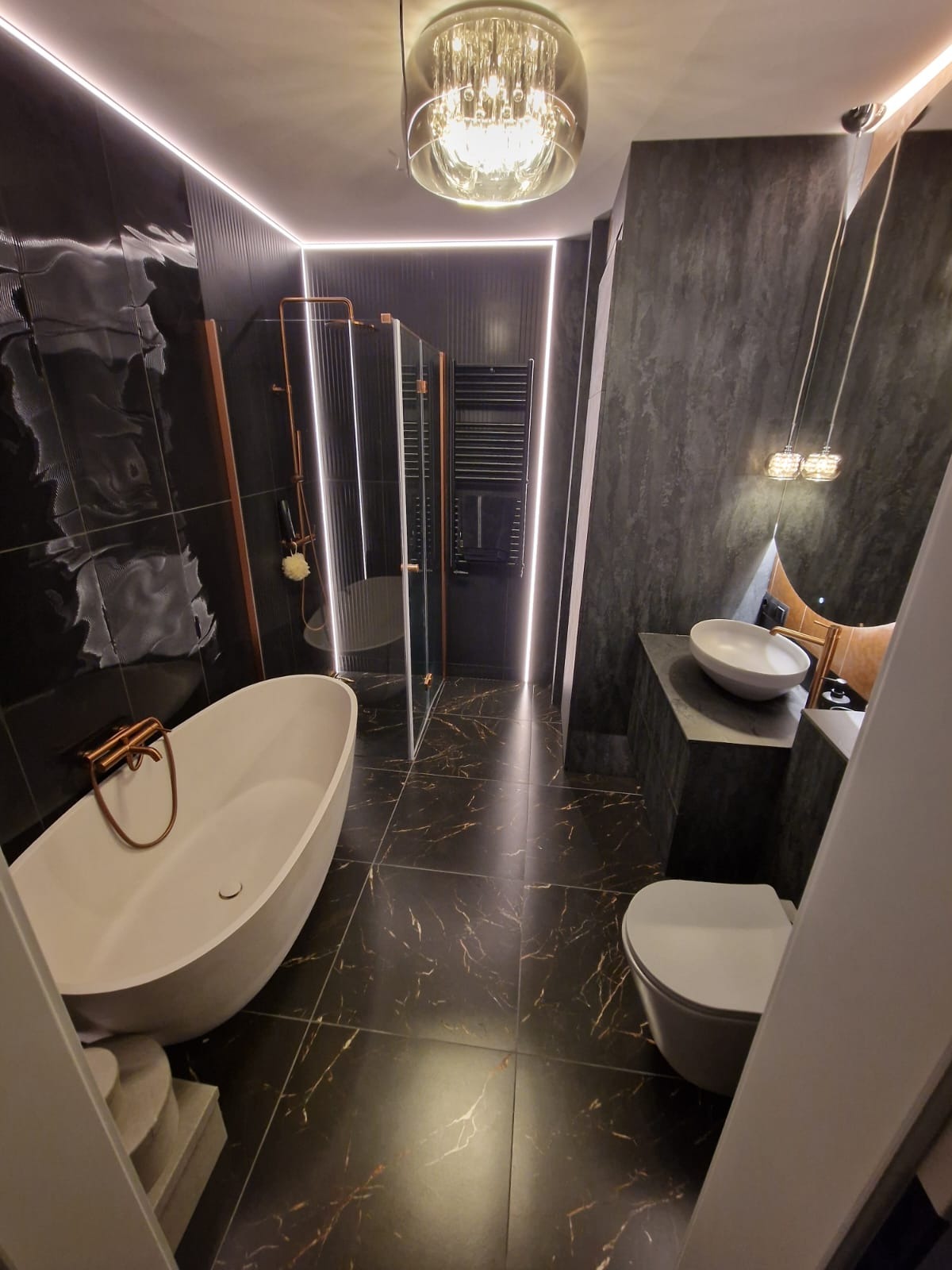 Elegancka łazienka w ciemnej tonacji z marmurowymi płytkami, wolnostojącą wanną i szklaną kabiną prysznicową. Oświetlenie LED podkreśla nowoczesny charakter wnętrza.