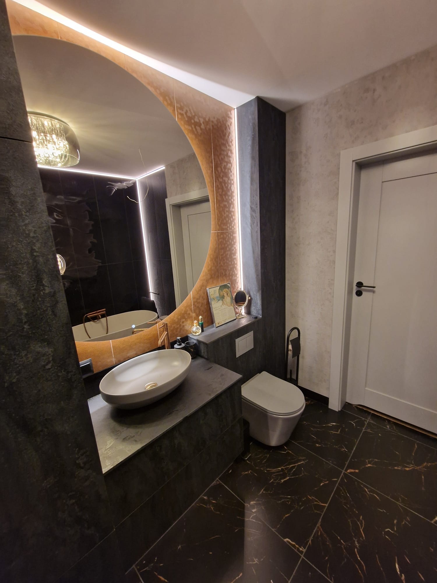Nowoczesna łazienka z owalną umywalką na blacie z ciemnego kamienia, duże lustro z podświetleniem i czarne płytki podłogowe z złotymi żyłami.