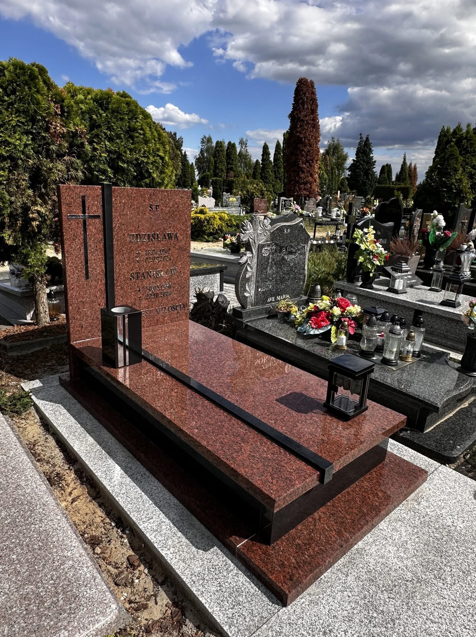 Granitowy nagrobek w odcieniach czerwieni z czarnymi akcentami, na tle cmentarza z zielenią i innymi nagrobkami. Widoczne napisy i krzyż.