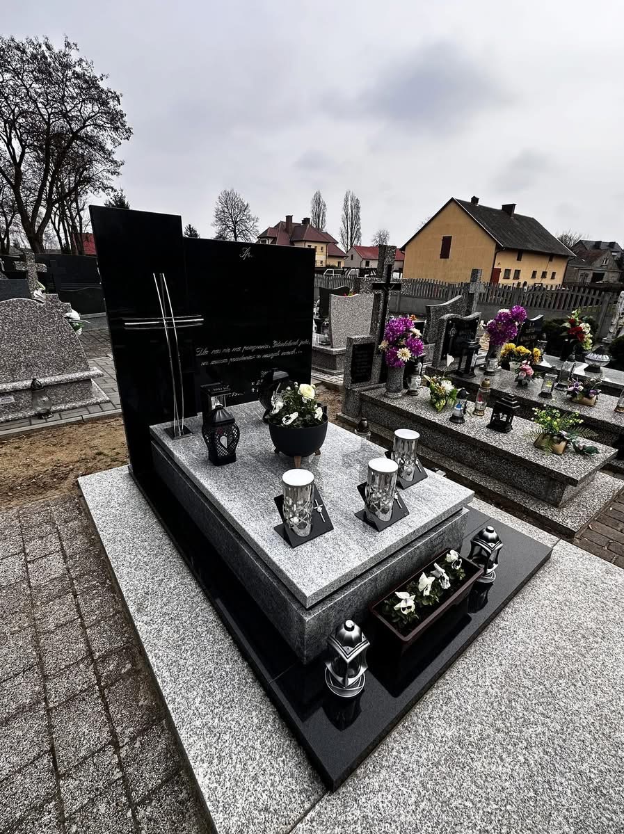 Czarny, nowoczesny nagrobek z jasnoszarego granitu, ozdobiony lampionami i kwiatami, na tle cmentarza z domami w oddali. Ujęcie z perspektywy.