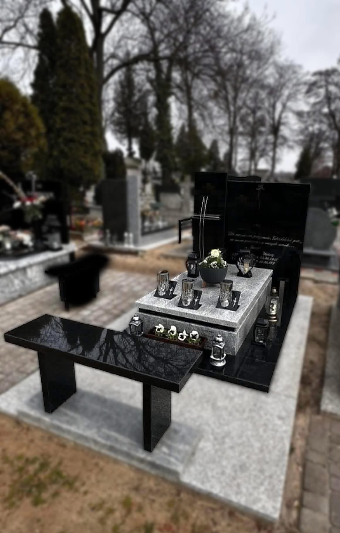 Czarno-szary nagrobek z ławką na cmentarzu, z kwiatami i lampionami. Polerowany kamień odbija drzewa. W tle inne nagrobki i drzewa bez liści.