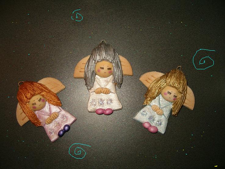Trzy ręcznie robione figurki aniołków z modeliny, z drewnianymi skrzydłami i różnymi kolorami włosów (złote, srebrne, rude), ułożone na ciemnym tle.