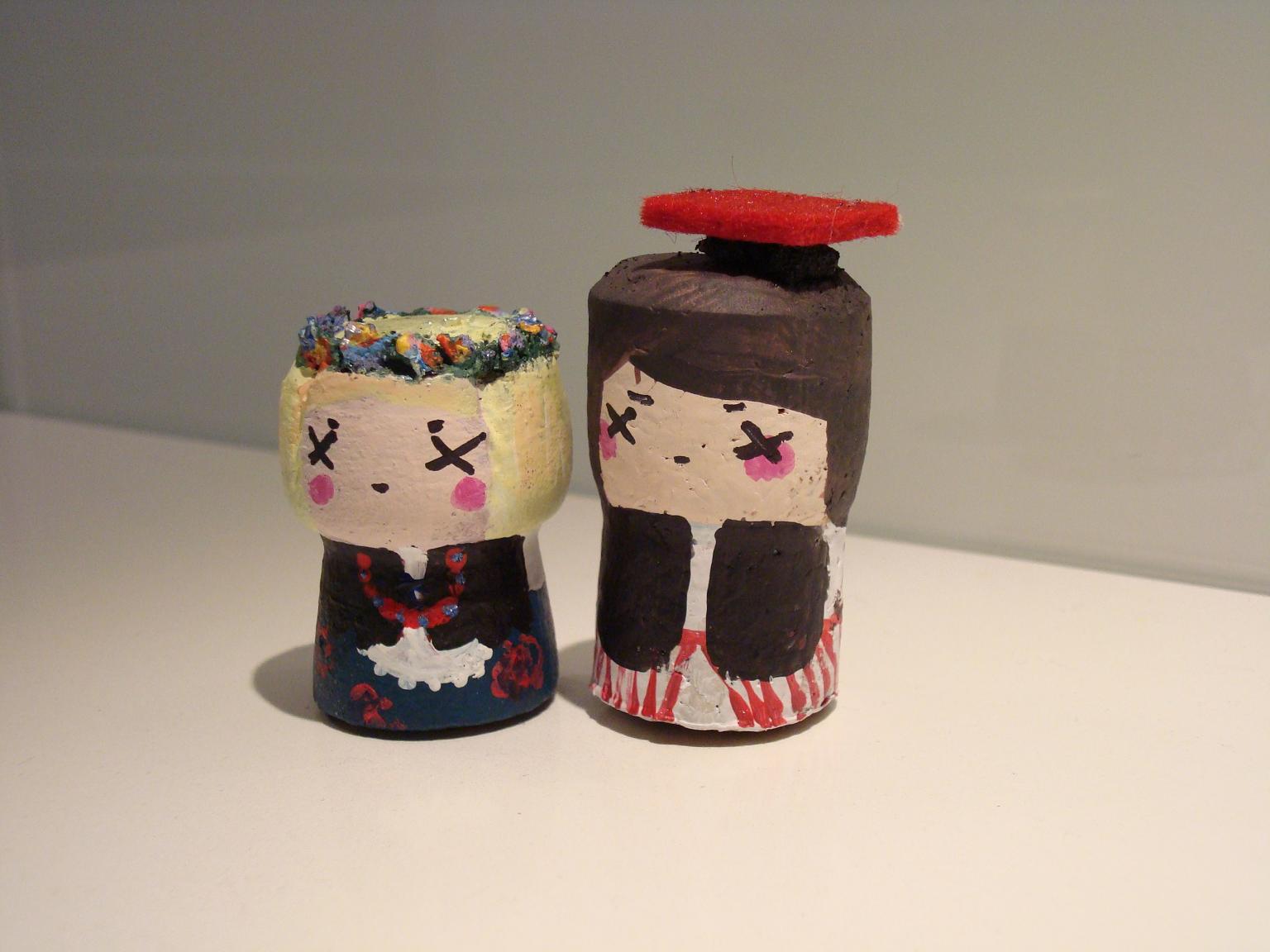 Ręcznie malowane drewniane figurki aniołków z charakterystycznymi znakami 'X' zamiast oczu, jedna z wiankiem z suszonych kwiatów, druga z czerwoną filcową czapką.