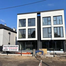 Montek - Nowoczesny, dwukondygnacyjny budynek z dużymi oknami i płaskim dachem w trakcie budowy. Widoczny baner z ofertą usług ogólnobudowlanych i materiały budowlane.