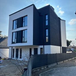 Montek - Nowoczesny, trzypiętrowy dom z czarno-białą elewacją, widoczny z perspektywy ulicy. Minimalistyczny design, duże okna, ogrodzenie z siatki. Częściowo zagospodarowany teren.