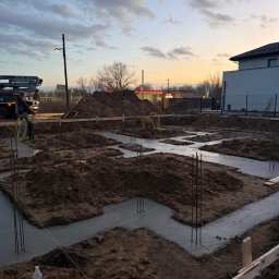 Montek - Zalane betonem fundamenty domu jednorodzinnego, widoczne wystające pręty zbrojeniowe i drewniane szalunki. W tle kopiec ziemi i budynek w trakcie budowy.