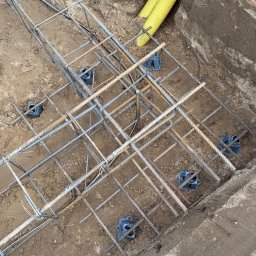 Montek - Zbrojenie fundamentów w wykopie, z użyciem dystansów z tworzywa. Żółta rura w tle. Widok z góry na przygotowany szkielet z prętów stalowych.