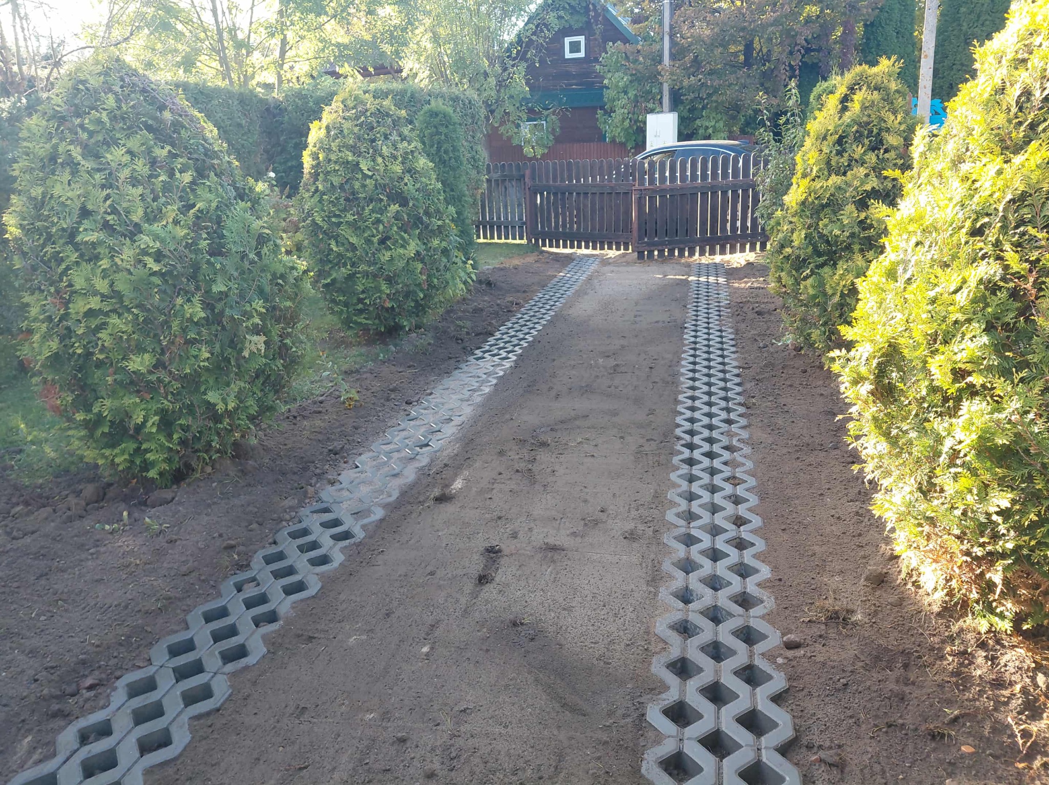 Ścieżka z ażurowych płyt betonowych prowadząca do drewnianej bramy, otoczona tujami. Widok na dom z ciemnego drewna w tle, w słoneczny dzień.