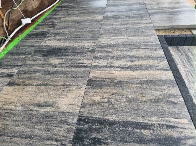 Ułożona kostka brukowa w odcieniach szarości i beżu, z widocznymi liniami podziału i fragmentem obrzeża. Widoczne kable przy ścianie.