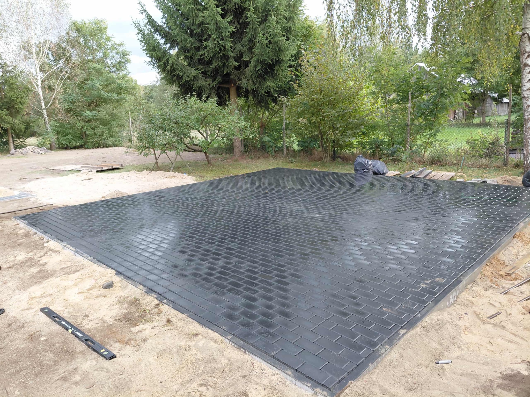 Ułożona, czarna kostka brukowa na piaskowym podłożu, otoczona zielenią drzew. Widoczny poziomica i worki na śmieci w tle. Świeżo położona nawierzchnia.