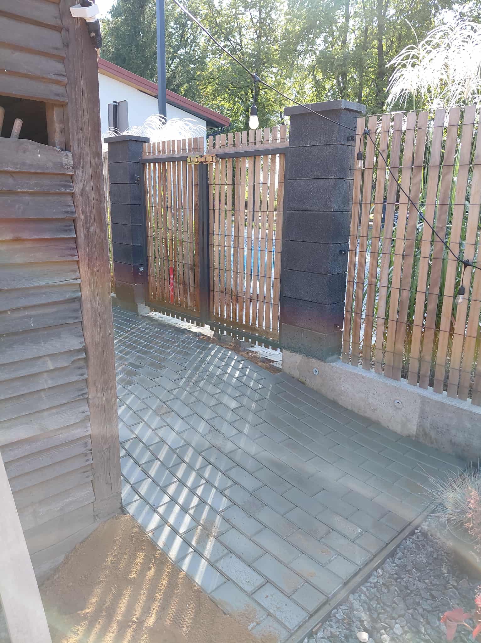 Świeżo ułożona, mokra kostka brukowa w odcieniach szarości, prowadząca do drewnianej furtki z elementami betonu. Widoczne oświetlenie zewnętrzne i fragment drewnianej konstrukcji.