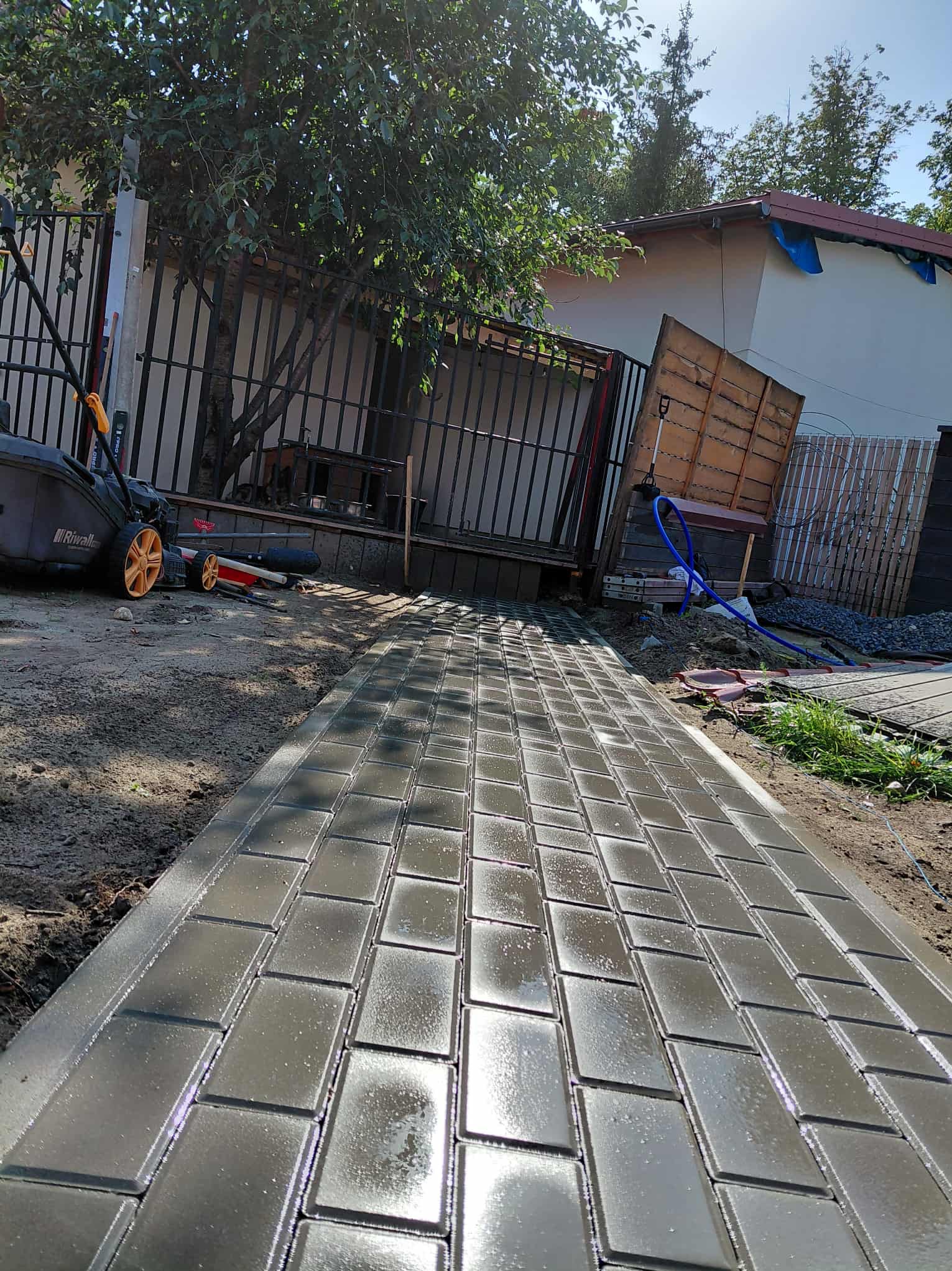 Świeżo ułożona, mokra kostka brukowa w odcieniach szarości, prowadząca w kierunku metalowej bramy i zieleni. W tle widoczny budynek i sprzęt ogrodniczy.