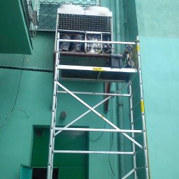 Bartosz - Klimatyzator Fujitsu na rusztowaniu przed budynkiem o zielonej elewacji. Widoczne otwarte drzwi i okno z kratą. Prace serwisowe w toku.