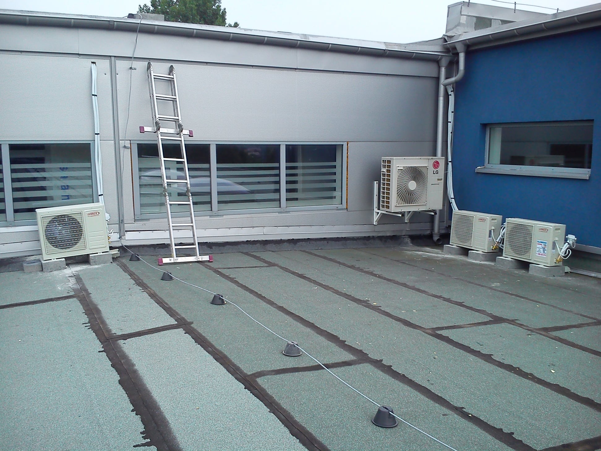 Instalacja klimatyzacji na dachu budynku z drabiny. Widoczne jednostki zewnętrzne klimatyzatorów LG i Lennox, kable, elementy montażowe i fragment elewacji.