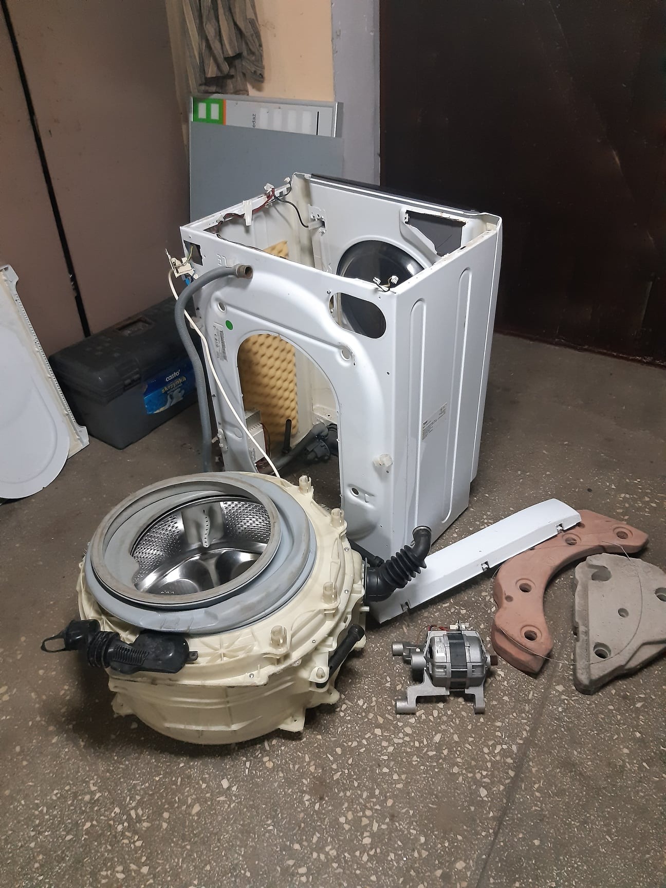 Rozłożona pralka automatyczna z widocznymi elementami: bęben, obudowa, silnik i inne części na podłodze. Widoczne ślady użytkowania i demontażu.