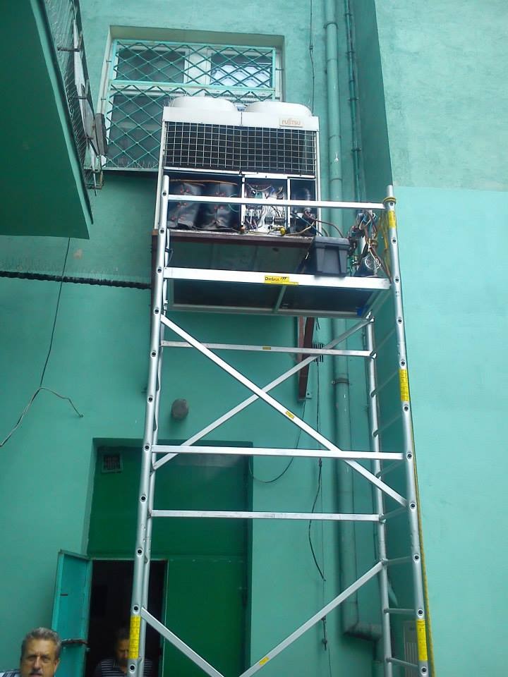 Klimatyzator Fujitsu na rusztowaniu przed budynkiem o zielonej elewacji. Widoczne otwarte drzwi i okno z kratą. Prace serwisowe w toku.